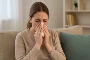 Young woman with allergies sitting on beige sofa holding tissue to nose, illustrating couch-related allergy symptoms
